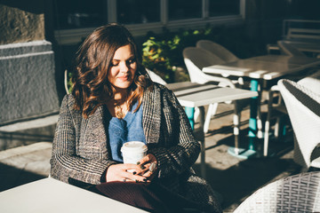 Young lady in coat holding cup of aromatic coffee in the city cafe.