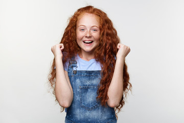 Wall Mural - Portrait of happy cute freckles little girl with ginger hair, with raised fists up, won a box of chocolates, stands over white wall and broadly smiling.