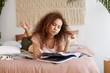 © timtimphoto - Young thinking african american woman with curly hair, lies on the bed, reads an article in a magazine in a concentrated manner.