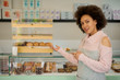 © chika_milan - Beautiful smiling mixed race female employee in apron standing in front of counter and showing fresh pastry.