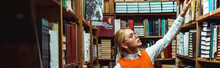 Panoramic Shot Of Pretty And Blonde Woman In Glasses Holding Book In Library