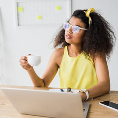 Wall Mural - thoughtful african american girl in glasses holding cup near laptop