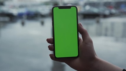 Los Angeles, California - 5 April 2019: Woman hand holding use touch vertically smart phone cellphone with green screen background blurred rain parking cars app people outside rain trees raindrops
