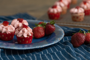 Homemade mini strawberry cupcakes in red and white paper liners.  Some isolated on blue plate with fresh strawberries.  Some cooling on rack in background.  Blue kitchen towel on wood table.