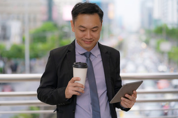 Wall Mural - Asian Businessman standing and holding digital tablet with business office buildings in the city background