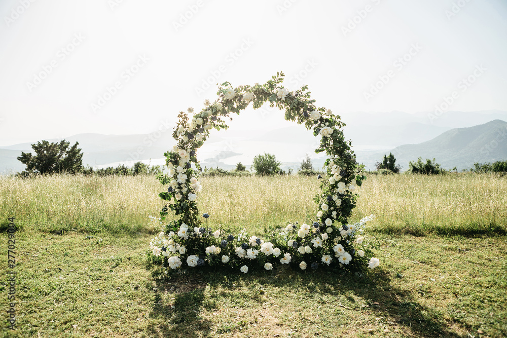 Beautiful wedding arch of flowers or white roses in a field or meadow. Preparation for a wedding event. - obrazy, fototapety, plakaty 
