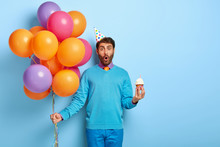 Surprised Young Man Celebrates Birthday, Holds Muffin With Burning Candle, Bunch Of Colored Air Balloons, Shocked With Unexpected News, Enjoys Party. Blue Colors. Time For Fun And Entertainment