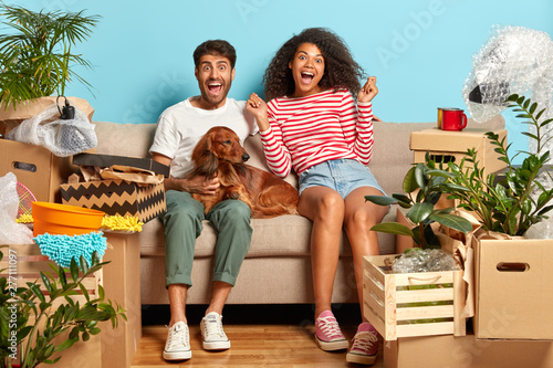 Overjoyed first time home buyers enjoy relocation, being tenant of new apartment, relaxes before unpacking boxes, sit on comfortable sofa in empty room with blue wallpapers, dog between them