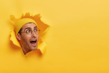 Overwhelmed Young Guy Keeps Head Through Torn Hole In Yellow Background, Keeps Jaw Dropped, Gazes Aside On Free Space, Notices Something Surprising, Wears Hat And Big Round Glasses. Reaction
