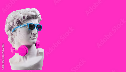 Statue. Earphone on a pink background. Gypsum statue of David\'s head. Creative. Plaster statue of David\'s head in blue sunglasses. Minimal concept art.