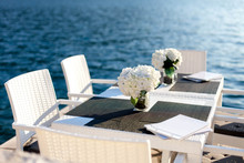Seafood Restaurant With White Furniture By Blue Water. Beautiful Serving With Hydrangea Flowers. Romantic Dinner Or Lunch Outdoors. Table And Chairs On Coastline.