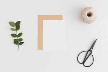 Sticker - Top view of a white card mockup and envelope with twine, scissors and green branch on a white table.