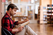 Casual Young Man Reading Book While Sitting By Bookshelf
