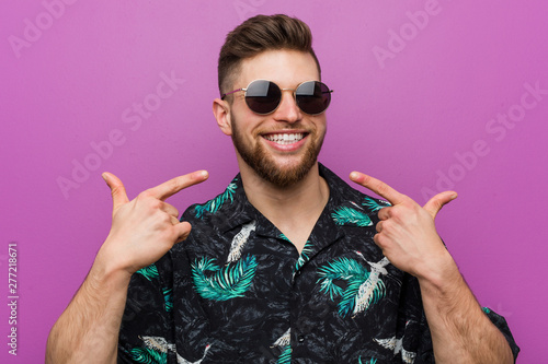 Fotomural Young man wearing a vacation look smiles, pointing fingers at mouth