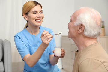 Wall Mural - Nurse feeding elderly man with yogurt indoors. Assisting senior people