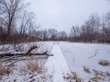 A Snow Covered Wood Plank And Steel Pier Sticking Out Into A Frozen Pond With Bare Tree Branches And Dead Vegetation And Weeds Framing The Shot In Rural Wisconsin With Cloudy Sky Above.