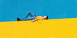 © Rafa Fernandez - Front view of a young boy wearing casual clothes lying on a yellow fence against a blue wall while using a mobile phone to listening music by headphones