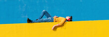 Front View Of A Young Boy Wearing Casual Clothes Lying On A Yellow Fence Against A Blue Wall While Using A Mobile Phone To Listening Music By Headphones
