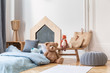 Stylish kid's bedroom in tenement house. Grey pouf on the parquet, wooden table with bags and toys, bed with blue bedding.