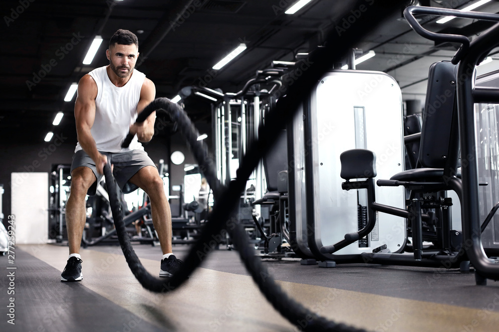 Athletic young man with battle rope doing exercise in functional training fitness gym. - obrazy, fototapety, plakaty 