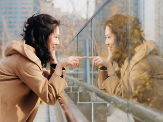 Beautiful Chinese girl with her mirror image in glass. Outdoor fashion portrait of glamour young Chinese cheerful stylish lady, emotions, people, beauty and lifestyle concept.