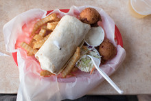 Spicy Chicken Wrap Resting On Crinkle Cut French Fries With A Side Of Organic Ranch Dip, Creamy Coleslaw Made From Local Produce, And Deep-fried Hush Puppies, All In A Red Plastic Basket