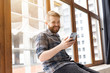 © satura_ - Cute young positive guy hipster with a beard is sitting on the windowsill by the big window and talking on the video communication using a smartphone and wireless high-speed Internet.
