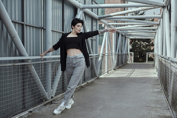 Raven haired female with tattoos is posing on the iron bridge in Tbilisi. She wears jacket, white sneakers and in gray jeans, Sunny weather with no clouds. Lady is sexy and playful. Luxury looking.