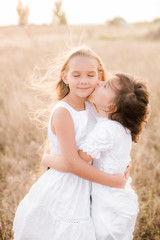 Cute little girls sisters with blond  hair in a summer field at sunset in white dresses with a straw hat