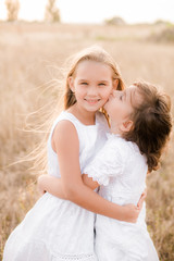 Cute little girls sisters with blond  hair in a summer field at sunset in white dresses with a straw hat