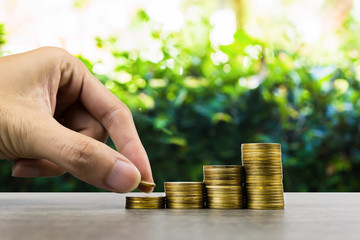 Long-term investment or making money with the right concepts. A business man hand putting on stack of coins on a wooden table with green nature as background. Depicts a standing and stable investment.