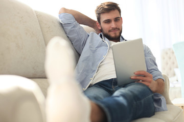 Sticker - Handsome man is using a digital tablet and smiling while resting on couch at home.