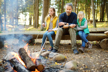 Wall Mural - Cute little sisters and their father roasting marshmallows on sticks at bonfire. Children having fun at camp fire. Camping with kids in fall forest.