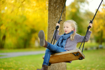 Wall Mural - Cute young girl having fun on a swing in sunny autumn park. Family weekend in a city.