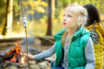 Wall Mural - Cute young sisters roasting marshmallows on stick at bonfire. Children having fun at camp fire. Camping with children in fall forest.