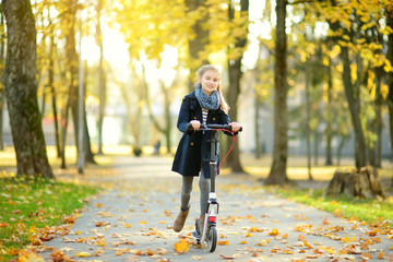 Wall Mural - Adorable young girl riding her scooter in a city park on sunny autumn evening. Pretty preteen child riding a roller.