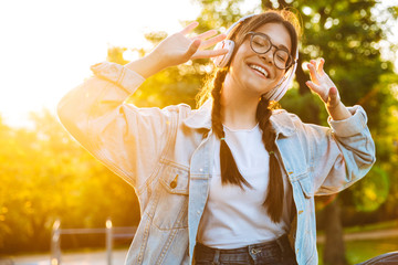 Wall Mural - Pleased happy young teenage girl student sitting outdoors in beautiful green park listening music with headphones.