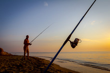 Man Is Fishing With Several Rods On The Beach In Sunrise Morning