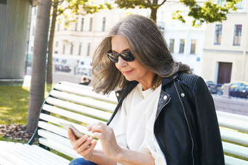 Wall Mural - Outdoor picture of busy modern middle aged woman in stylish black sunglasses and leather jacket messaging online using smart phone, sitting on empty street in the morning. Technology and communication