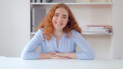 Poster - Successful businesswoman sitting at white desk wearing casual dress in contemporary office. Attractive woman posing looking at the camera. Beautiful lady with curly red hair and freckles on young face