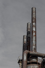 Wall Mural - Trio of rusted and riveted metal smokestacks against a dark grey sky, copy space, vertical aspect