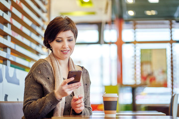 woman in a cafe with a paper cup of coffee. brown sweater and scarf. sits by the table. talking on the phone