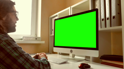 Leinwandbilder - man sitting at workplace paying game on computer in small office. businessman with beard looking on display with green screen. employee in startup company typing on keyboard use pc mouse
