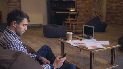 Poster - Attractive man in a plaid shirt check the data on the phone and on the printed document. On the table is a laptop and a cup of coffee costs.
