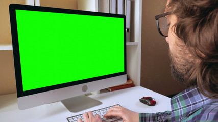 Leinwandbilder - businessman sitting at workplace in small office. man with beard looking on display computer with green screen. employee in startup company typing on keyboard