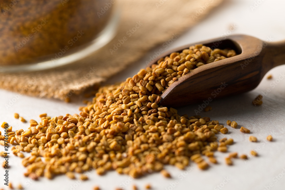 Dried, raw fenugreek seed in wooden scoop with storage glass jar on white wooden table background
