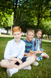 © Seventyfour - Three smiling teenage friends sitting on grass in park in summer under shadow of big high trees