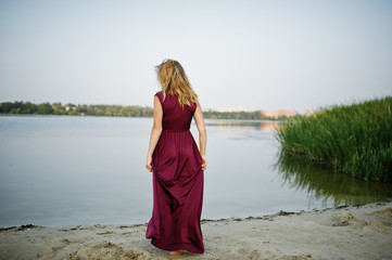 Wall Mural - Back of blonde sensual barefoot woman in red marsala dress posing against lake with reeds.