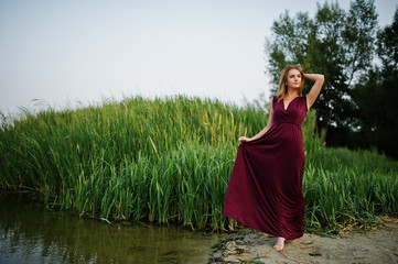 Wall Mural - Blonde sensual barefoot woman in red marsala dress posing against lake with reeds.