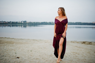 Wall Mural - Blonde sensual barefoot woman in red marsala dress posing against lake on sand.